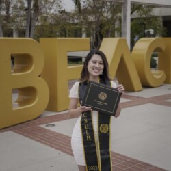 portrait of Pia holding a diploma in front of a large "BEACH" sign